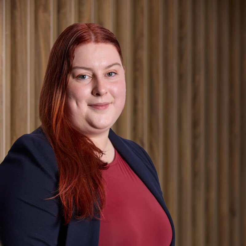 Headshot of Lily wearing a red top and blazer on a brown wooden background